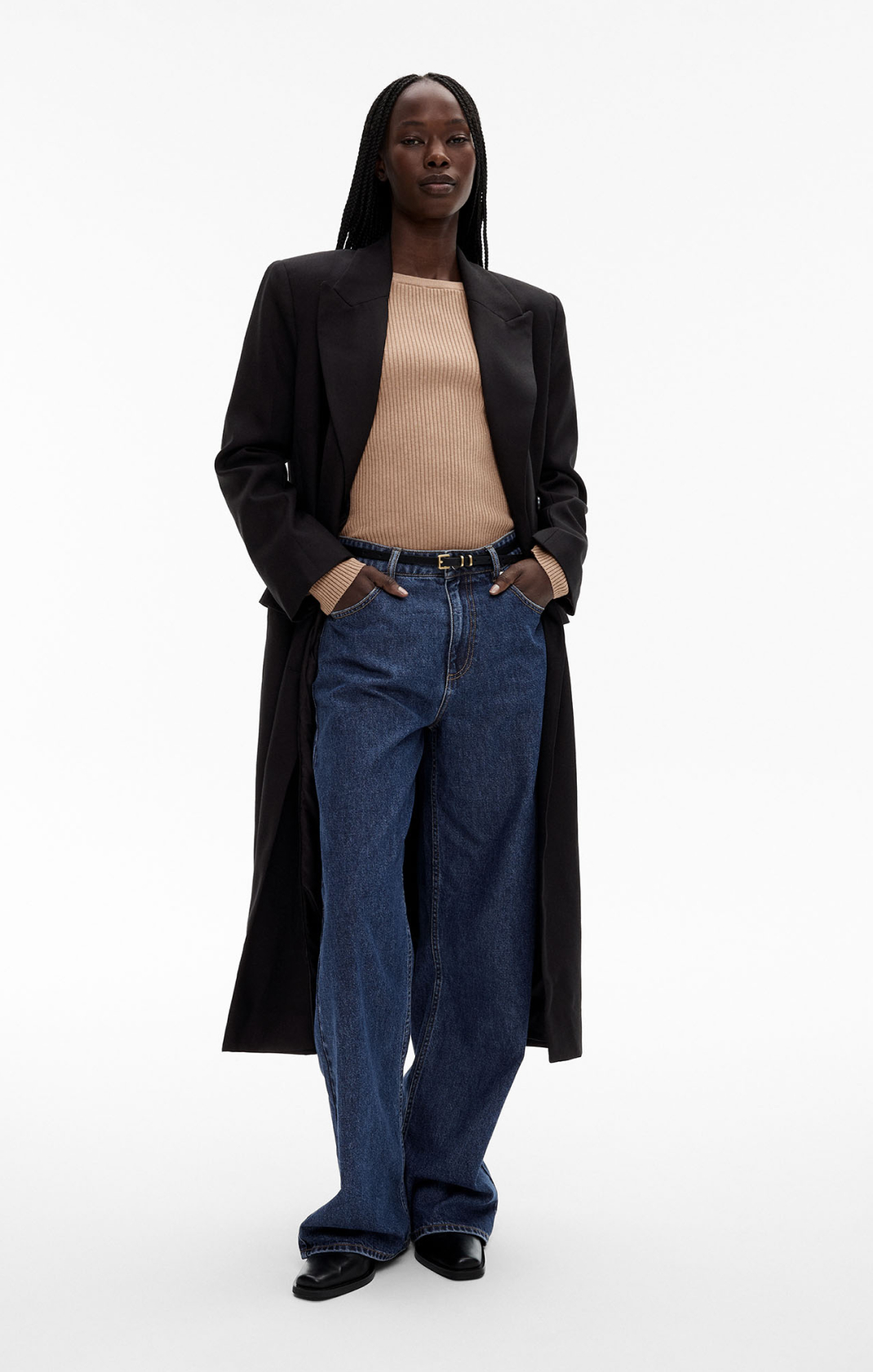 Woman in tan long sleeve knit, black coat, indigo denim jeans and black leather belt
